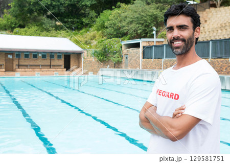 Male lifeguard standing at pool edge wearing T shirt monitoring lanes at outdoor pool, copy space 129581751