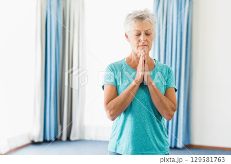 Senior woman standing next to tall curtains in home studio meditating in teal workout shirt Senior woman standing next to tall curtains in home studio meditating in teal workout shirt 129581763