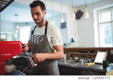 Male barista steaming milk with metal pitcher behind espresso machine at coffee shop counter 129581771