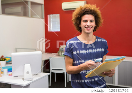 Curly-haired man standing in office holding smartphone and manila folders by monitor, copy space 129581792