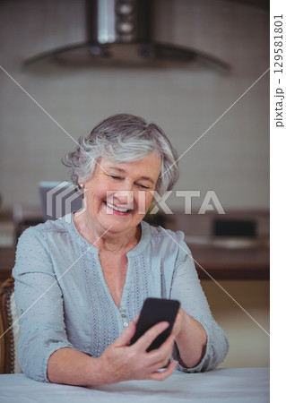 Senior woman holding smartphone and smiling at home kitchen table with laptop, hood vent, items 129581801