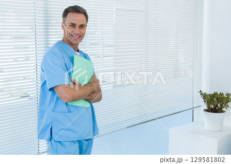 Middle-aged male wearing scrubs standing in office holding green folder beside plant, copy space 129581802