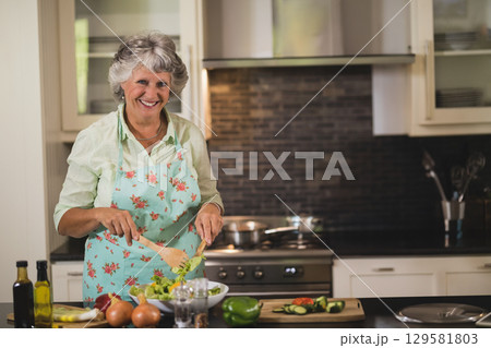 Senior woman tossing salad in white bowl with wooden spoon at kitchen island with sliced cucumbers 129581803
