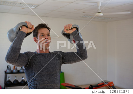 Man holding two kettlebells at shoulder height in fitness studio with rack, mat and bench 129581875