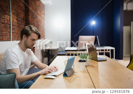 Man working on tablet and keyboard at wooden communal table in coworking space with coffee cup 129581876