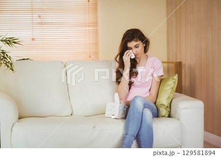 Woman sitting on sofa in living room holding tissue wiping tears beside tissue box Woman sitting on sofa in living room holding tissue wiping tears beside tissue box 129581894