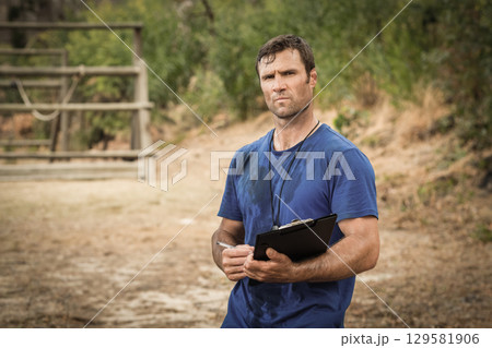 Man wearing blue T-shirt holding clipboard, pen and whistle inspecting wooden obstacle course Man wearing blue T-shirt holding clipboard, pen and whistle inspecting wooden obstacle course 129581906