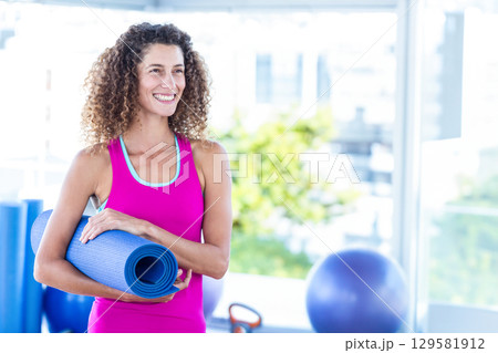 Woman holding rolled blue yoga mat at gym, wearing fuchsia tank top and leggings with kettlebell Woman holding rolled blue yoga mat at gym, wearing fuchsia tank top and leggings with kettlebell 129581912