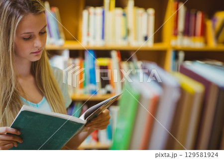 Teenage girl reading open book in library aisle with bookshelves and blurred books, copy space 129581924