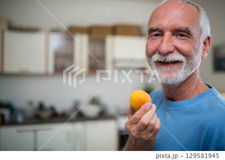Senior man standing in kitchen holding yellow apricot near mouth, wearing blue t-shirt, copy space 129581945
