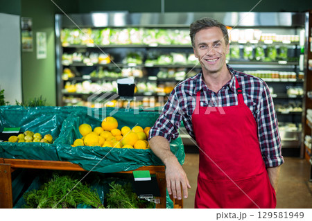 Middle-aged man in apron leaning on bin at supermarket produce aisle showing lemons with price sign 129581949