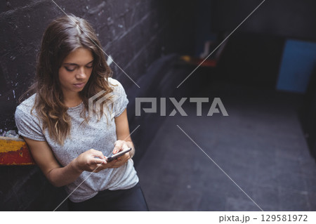 Woman mid-20s leaning against painted wall in urban alley holding smartphone near ledge, copy space Woman mid-20s leaning against painted wall in urban alley holding smartphone near ledge, copy space 129581972