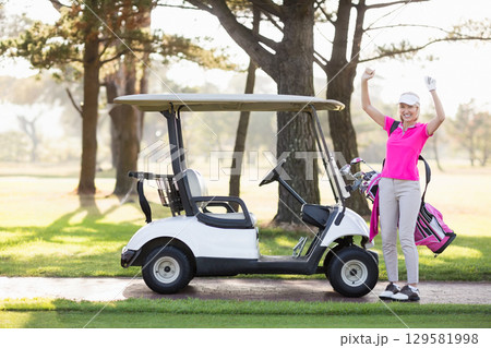 Woman in pink polo celebrating beside cart and clubs on golf course path, copy space 129581998