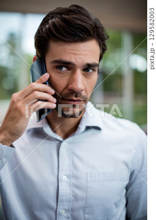 Middle-aged man standing near windows in modern office holding smartphone to ear wearing blue shirt 129582003