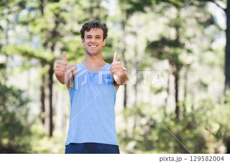 Smiling man extending arms with thumbs up in sunlit forest clearing wearing athletic wear 129582004