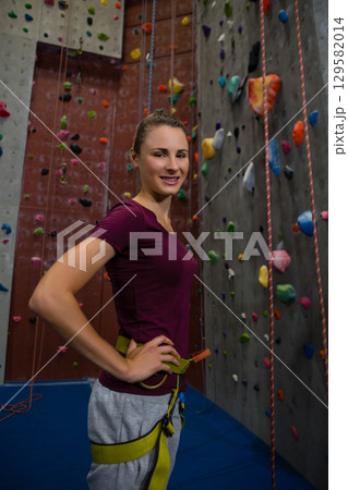 Female climber standing at climbing gym wearing yellow harness near colorful holds and ropes Female climber standing at climbing gym wearing yellow harness near colorful holds and ropes 129582014