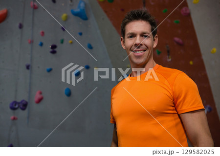 Mid adult man posing by climbing wall wearing orange shirt with holds and rope, copy space 129582025