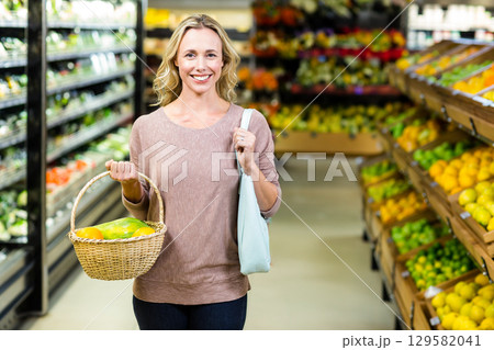 Woman in her 30s shopping in produce aisle holding basket of bell peppers and tote bag 129582041