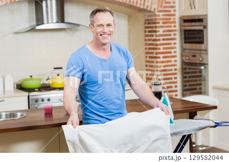Senior man ironing white shirt with steam iron on ironing board in home kitchen with pots Senior man ironing white shirt with steam iron on ironing board in home kitchen with pots 129582044