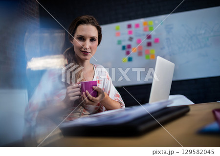 Female worker sitting at desk in meeting room, holding pink mug near silver laptop under whiteboard 129582045