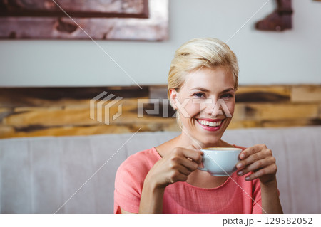 Woman wearing coral blouse smiling and holding white ceramic cup on café bench by wood-paneled wall Woman wearing coral blouse smiling and holding white ceramic cup on café bench by wood-paneled wall 129582052