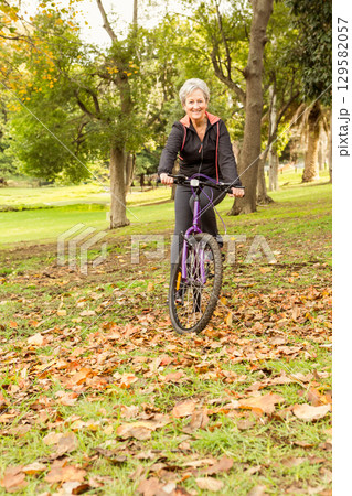 Senior woman cycling through park in autumn on purple bicycle wearing black jacket with earbuds 129582057