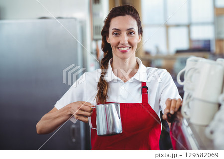 Female barista in red apron, holding frothing pitcher and using steam wand at coffee shop counter 129582069