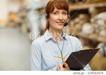 Mid adult woman standing in warehouse aisle wearing headset and holding clipboard near boxes 129582074