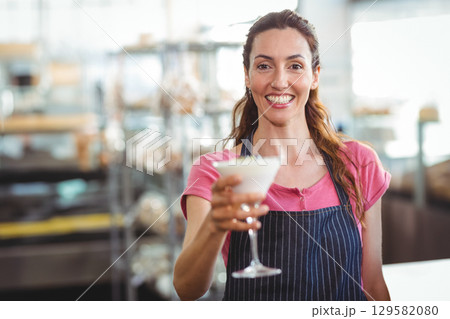 Mid adult female bartender in dark striped apron extending frothy martini glass at kitchen shelves 129582080