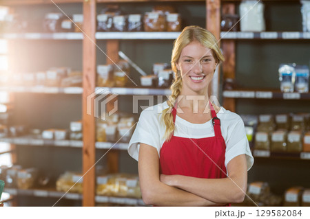 Female clerk standing with arms crossed in shop by wooden shelves with bulk jars and scanner Female clerk standing with arms crossed in shop by wooden shelves with bulk jars and scanner 129582084