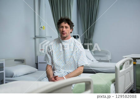 Male patient wearing hospital gown sitting on bed in ward beside IV stand and privacy curtains 129582088