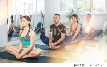 Concentrated young woman performing seated bound angle pose or butterfly pose Baddha Konasana during yoga class with group multinational people in fitness studio 129582126