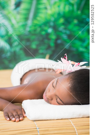 African American woman reclining on bamboo mat spa table outside wearing pink flower and towel African American woman reclining on bamboo mat spa table outside wearing pink flower and towel 129582525