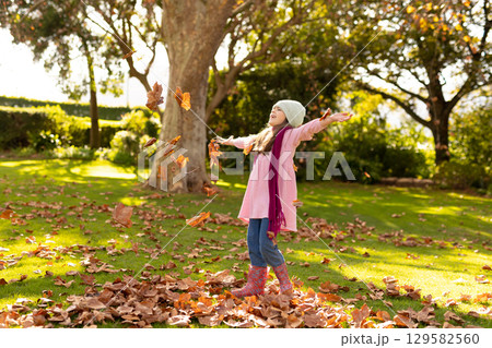 Female child tossing colorful leaves into air on sunlit grassy lawn wearing beanie and rain boots 129582560