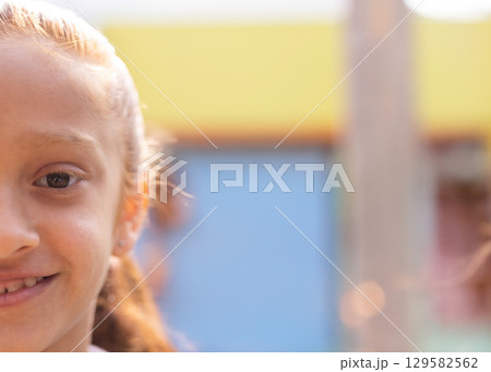Girl smiling and gazing at camera by wooden post near pastel walls in courtyard, copy space 129582562