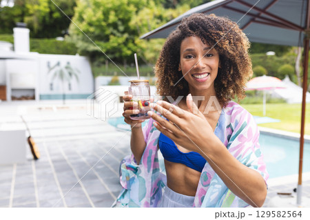 African American woman standing on pool deck holding fruit infused jar under umbrella beside guitar 129582564