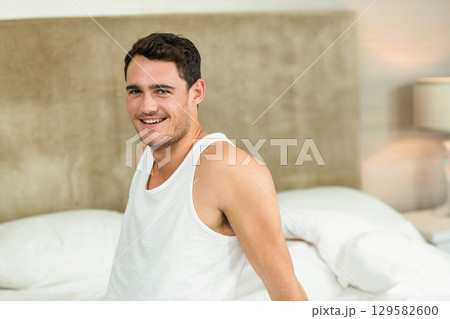 Male sitting on bed smiling under lamp with white sheets and beige headboard leaning back Male sitting on bed smiling under lamp with white sheets and beige headboard leaning back 129582600