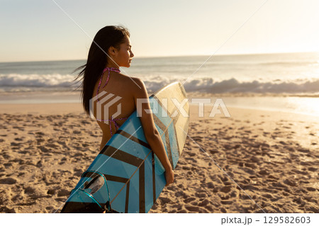 Asian woman in her twenties wearing bikini holding blue-and-black striped surfboard at water edge 129582603