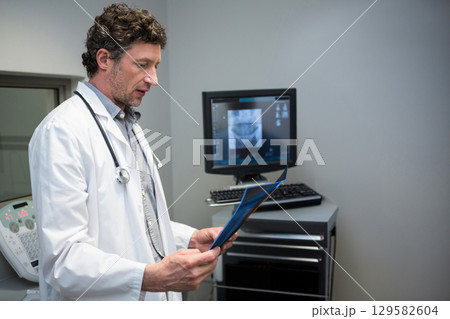Middle-aged doctor holding blue x-ray folder with stethoscope and examining monitor in clinic Middle-aged doctor holding blue x-ray folder with stethoscope and examining monitor in clinic 129582604