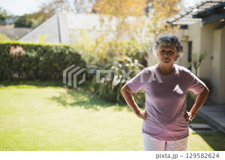 Senior woman standing with hands on hips in suburban backyard by hedge and deck, copy space 129582624