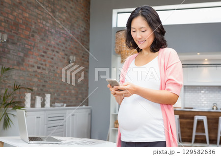 Pregnant Asian woman standing in kitchen smiling and checking smartphone by open laptop, copy space 129582636
