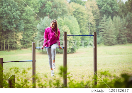 Woman in pink windbreaker performing hanging leg raises on metal exercise frame in park clearing Woman in pink windbreaker performing hanging leg raises on metal exercise frame in park clearing 129582637