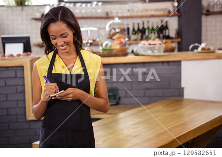 African American woman in apron standing behind cafe counter writing on notepad beside pastries African American woman in apron standing behind cafe counter writing on notepad beside pastries 129582651