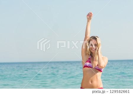 Adult woman standing at shoreline of turquoise sea raising arm overhead in pink bikini, copy space 129582661