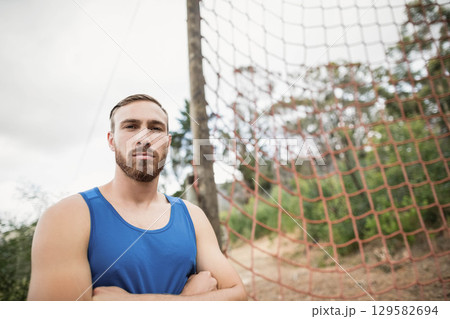 Male athlete wearing blue top standing with arms crossed on outdoor obstacle course with rope net Male athlete wearing blue top standing with arms crossed on outdoor obstacle course with rope net 129582694