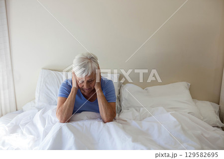 Senior woman sitting on bed wearing light blue shirt holding hands to temples on white comforter Senior woman sitting on bed wearing light blue shirt holding hands to temples on white comforter 129582695