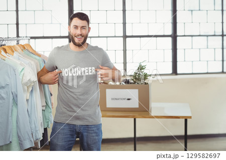 Male volunteer pointing at VOLUNTEER T-shirt while smiling by donation box in charity room Male volunteer pointing at VOLUNTEER T-shirt while smiling by donation box in charity room 129582697