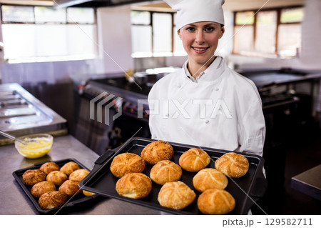 Female chef in chef coat and hat, holding baking tray with golden rolls in restaurant kitchen Female chef in chef coat and hat, holding baking tray with golden rolls in restaurant kitchen 129582711