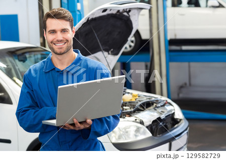 Male mechanic in blue coveralls holding laptop while diagnosing sedan engine in garage bay 129582729