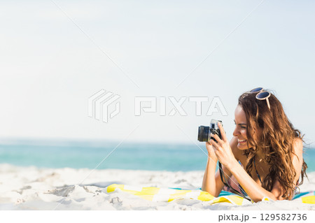 Woman in bikini lying on beach towel shooting blue ocean holding camera and sunglasses, copy space Woman in bikini lying on beach towel shooting blue ocean holding camera and sunglasses, copy space 129582736